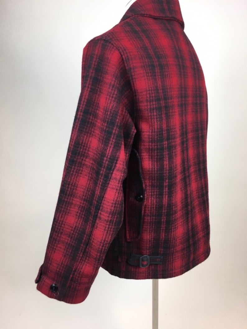 May include: A red and black plaid jacket with a button closure and a small buckle detail at the waist. The jacket has a classic collar and long sleeves. The fabric appears to be a thick wool or wool blend.