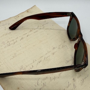May include: Vintage tortoiseshell sunglasses with green lenses, resting on aged paper with handwritten script. The frame has a classic, retro design. The glasses are a stylish accessory.