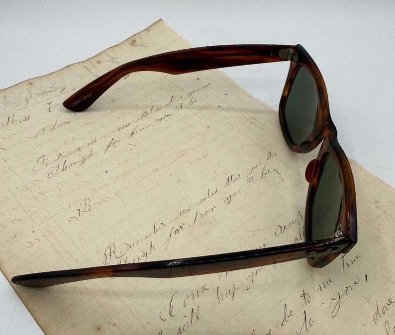May include: Vintage tortoiseshell sunglasses with green lenses, resting on aged paper with handwritten script. The frame has a classic, retro design. The glasses are a stylish accessory.