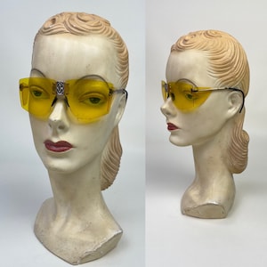 Puede incluir: Gafas de sol de color amarillo con montura plateada y un logotipo en el puente de la nariz. Las gafas de sol están en una cabeza de maniquí.