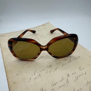 May include: Cat-eye shaped sunglasses in a brown tortoiseshell pattern, placed on a vintage handwritten letter. The sunglasses have a dark brown frame and tinted lenses. The letter features cursive handwriting.