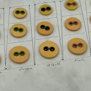 Puede incluir: Una colección de botones vintage en varios colores, incluyendo amarillo, marrón, verde, negro, rosa, azul y rojo. Cada botón tiene dos agujeros y está dispuesto en filas sobre una superficie blanca.