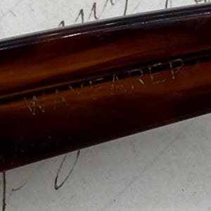 May include: Close-up of a brown Wayfarer frame. The text "WAYFARER" is etched into the frame. The frame is a dark, translucent brown colour. The background is a light colour with handwritten script.