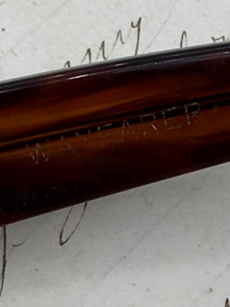 May include: Close-up of a brown Wayfarer frame. The text "WAYFARER" is etched into the frame. The frame is a dark, translucent brown colour. The background is a light colour with handwritten script.