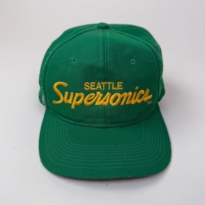 Może przedstawiać: Zielona czapka bejsbolowa z płaskim daszkiem. Czapka ma wyszyty napis "SEATTLE SuperSonics" w kolorze żółtym, klasyczne logo drużyny sportowej. Kapelusz jest wykonany z wytrzymałej tkaniny.