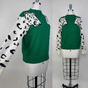 Può includere: Maglione verde con maniche bianche con macchie nere. Il maglione ha uno scollo tondo e un disegno a testa di leopardo sul petto. I polsini sono color crema.
