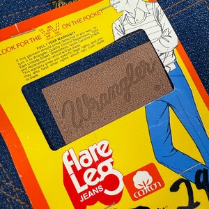 Pode incluir: Close-up de um par de jeans Wrangler azuis de perna larga. Os jeans t&ecirc;m um patch de couro castanho com o logotipo Wrangler. Uma etiqueta amarela com texto vermelho diz "Flare Leg Jeans" e "cotton".