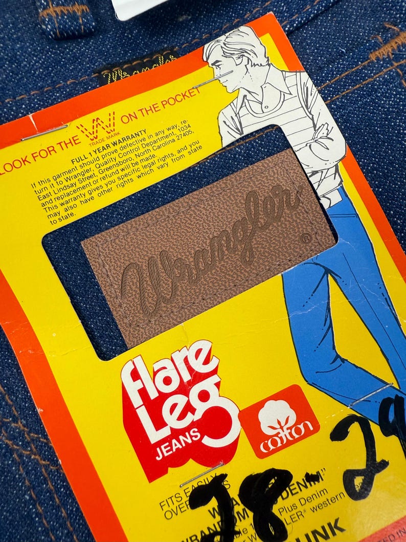 Pode incluir: Close-up de um par de jeans Wrangler azuis de perna larga. Os jeans t&ecirc;m um patch de couro castanho com o logotipo Wrangler. Uma etiqueta amarela com texto vermelho diz "Flare Leg Jeans" e "cotton".