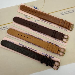 May include: Four watch straps in varying shades of brown leather, each attached to a light pink backing card. The straps feature metal buckles and are labeled with the text genuine top grain leather and watch strap, with Made in U.S.A. printed.