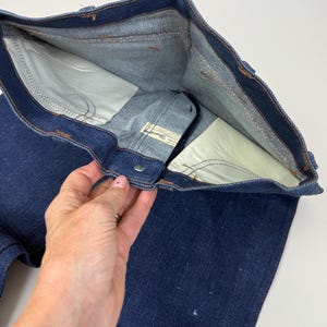 Pode incluir: Cal&ccedil;as de ganga azul escuro com fecho de bot&atilde;o e forro interior branco. As cal&ccedil;as de ganga est&atilde;o abertas, mostrando os bolsos internos e os detalhes das costuras. A etiqueta da marca &eacute; vis&iacute;vel. As cal&ccedil;as de ganga s&atilde;o feitas de ganga resistente.