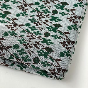 Op de afbeelding: Witte stof met een groen en bruin geometrisch patroon. De stof is dubbelgevouwen en het patroon is aan beide kanten zichtbaar.