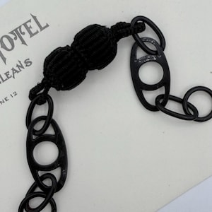 May include: Black chain accessory with oval links and a knotted detail. The accessory is on a white surface with the word "HOTEL" and "LEANS" visible in the background.