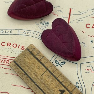 May include: Two dark purple heart-shaped buttons with a leaf design. The buttons are about 1 inch in diameter and are sitting on a piece of paper with a ruler.