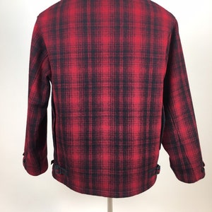 May include: A red and black plaid jacket with a classic collar and buttoned cuffs. The back of the jacket is visible, showcasing the plaid pattern and two adjustable straps near the hem. The jacket is made of a thick, warm material.