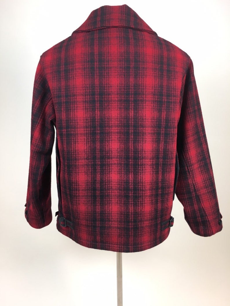 May include: A red and black plaid jacket with a classic collar and buttoned cuffs. The back of the jacket is visible, showcasing the plaid pattern and two adjustable straps near the hem. The jacket is made of a thick, warm material.