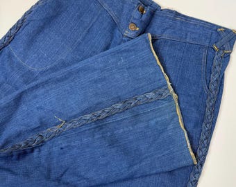 Può includere: Pantaloni di jeans blu con gamba larga e finiture a treccia decorative sui lati. I pantaloni hanno una vita alta con due bottoni e una piccola etichetta con la scritta "REAL WEST".