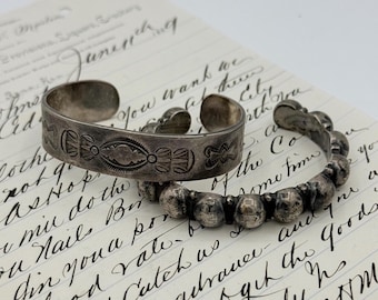May include: Two silver cuff bracelets. One bracelet is a wide band with stamped geometric patterns. The second bracelet is a narrow band composed of a series of connected silver spheres. Both items have a tarnished, metallic finish.