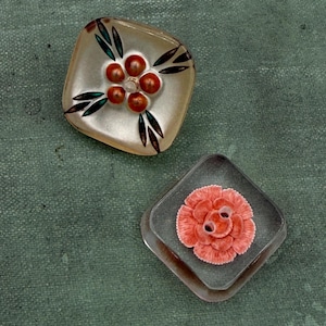 May include: Two square buttons with floral designs. One button is clear with a flower design of red berries and green leaves. The other button is gray with a coral-colored flower.