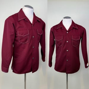 May include: A burgundy long-sleeve shirt with two chest pockets and a pointed collar. The shirt features white stitching along the collar, pockets, and seams. It has silver buttons and is displayed on a mannequin.