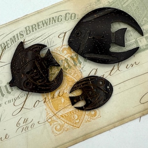 May include: Three dark brown, fish-shaped decorative objects. The fish are carved from a material with a speckled texture, possibly wood or coconut shell. They are arranged on a vintage document with text including "GEMIS BREWING CO" and "TWO CENTS".