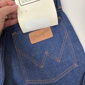 Puede incluir: Primer plano de unos vaqueros de mezclilla azul oscuro con un parche Wrangler de cuero marr&oacute;n en el bolsillo trasero. Los vaqueros tienen costuras naranjas y una etiqueta blanca con texto.