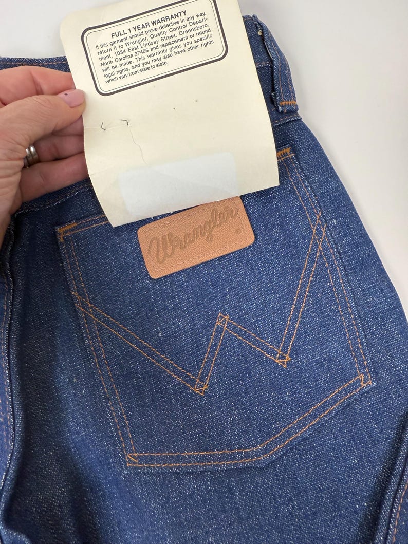 Puede incluir: Primer plano de unos vaqueros de mezclilla azul oscuro con un parche Wrangler de cuero marr&oacute;n en el bolsillo trasero. Los vaqueros tienen costuras naranjas y una etiqueta blanca con texto.