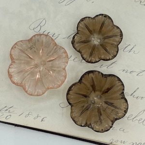 May include: Three flower-shaped glass buttons in shades of pink and brown. The buttons have a detailed petal design and small holes for sewing. They are arranged on a vintage paper background with handwritten text.