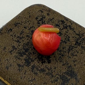 May include: A small, red, round object with a yellow ring on top, set against a dark, textured background. The object appears to be a decorative piece or a small sculpture, possibly made of glass or a similar material.