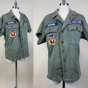 Può includere: Una camicia a maniche corte color verde oliva con bottoni e bordi delle maniche sfrangiati. La camicia presenta una targhetta blu con il nome ARANA, una toppa blu U.S. AIR FORCE e un emblema militare ricamato a colori con spada e ali.