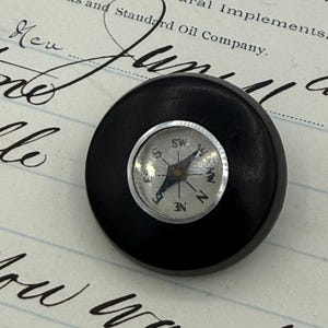 May include: A small, round, black compass with a silver-colored compass face. The compass needle points north. The compass is set against a background of old paper with handwritten text.