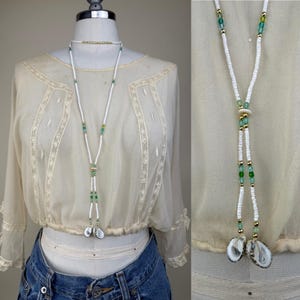 Può includere: Collana di perline bianche con accenti verdi e oro e due ciondoli a conchiglia. La collana è esposta su un manichino che indossa una camicetta color crema e jeans blu. La collana è uno stile lariat a forma di Y.