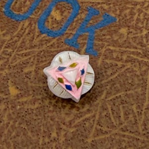 Könnte beinhalten: Ein kleiner, dekorativer Knopf mit einem rosa Dreieck-Design, akzentuiert mit blauen, grünen und gelben Details. Der Knopf befindet sich auf einer weißen Basis mit goldenen Akzenten. Der Hintergrund ist eine strukturierte braune Oberfläche.
