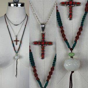 May include: Necklaces with cross pendants and decorative elements. One necklace features a silver cross pendant with red accents. Another necklace has a white stone pendant with a black tassel. The necklaces are displayed on a mannequin.
