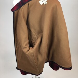 May include: A brown jacket with a red and black plaid collar and trim. The jacket has a brown interior lining and a black zipper. The Woolrich brand label is visible inside the jacket.