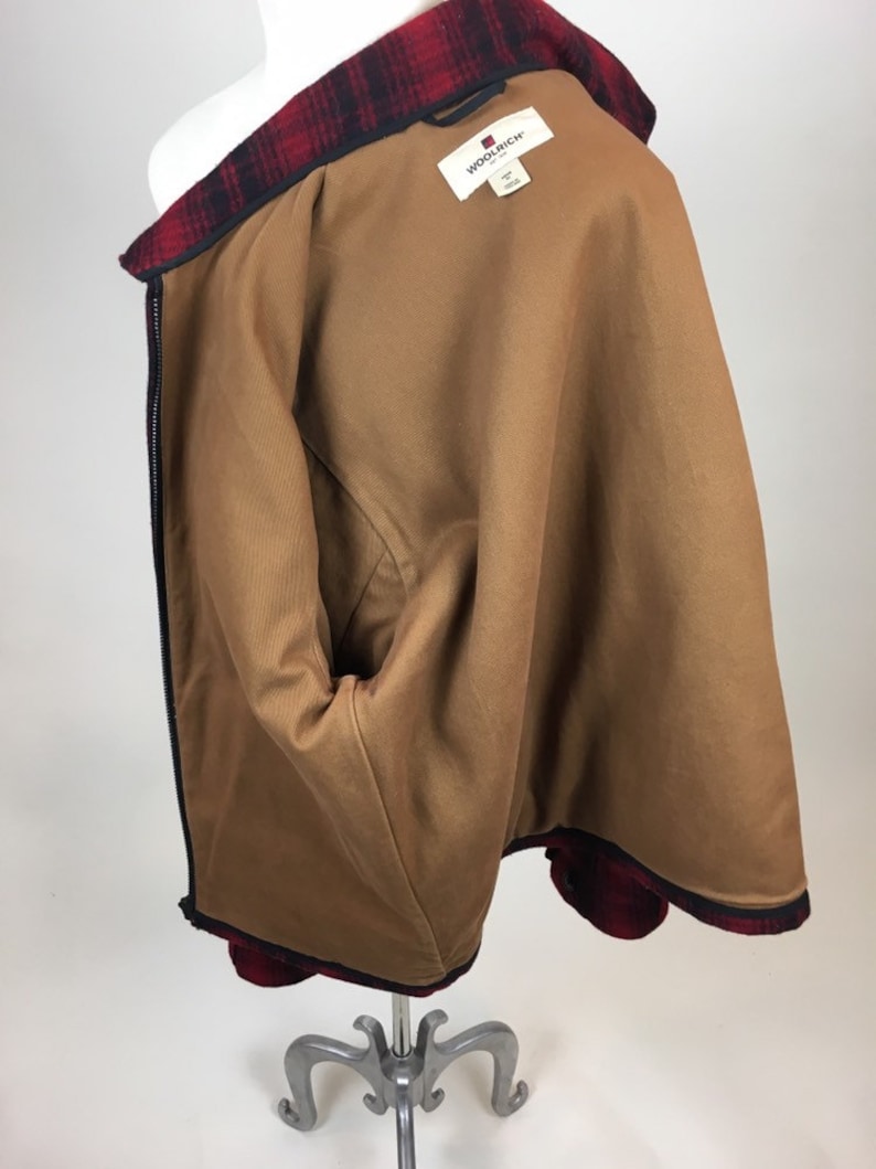 May include: A brown jacket with a red and black plaid collar and trim. The jacket has a brown interior lining and a black zipper. The Woolrich brand label is visible inside the jacket.