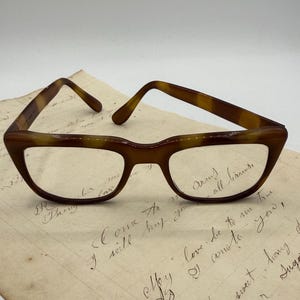 May include: Brown tortoiseshell eyeglasses with a cat-eye shape, resting on a vintage handwritten letter. The glasses have a classic design with clear lenses and a dark brown frame. The letter features cursive handwriting.