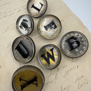 May include: A collection of vintage letterpress printing blocks. Each circular block features a different letter in black against a silver or gold background. The letters include L, E, F, U, W, B, and X. The blocks are arranged on a sheet of aged paper.