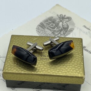 May include: A pair of cufflinks in a box. The cufflinks are made of silver and have a black and brown tortoiseshell design. The box is gold and has the text "Distinctive Creations 522 Fifth Ave Seattle" on it.