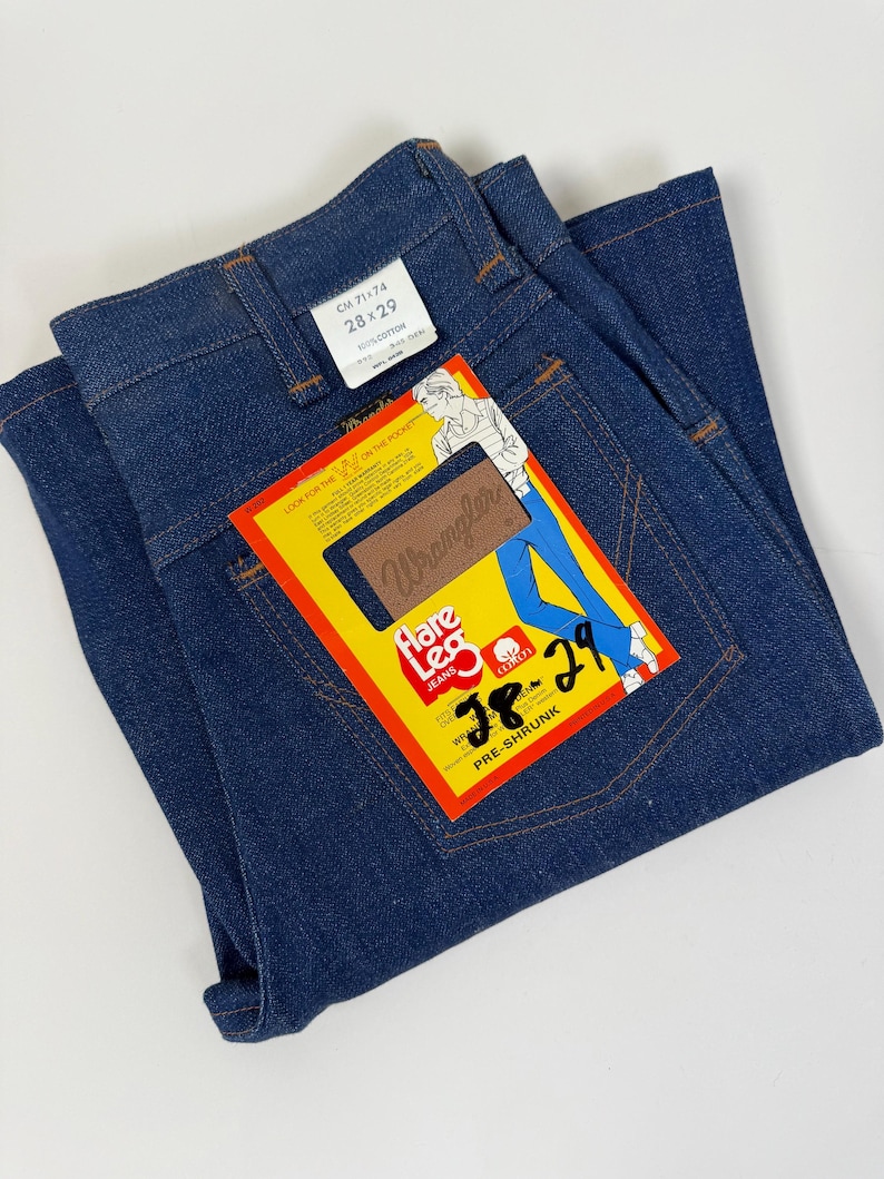 Pode incluir: Cal&ccedil;as de ganga azul escuro com uma etiqueta "Wrangler". A etiqueta amarela apresenta "Flare Leg Jeans" e o tamanho "28x29" (aproximadamente 71x74 cm). As cal&ccedil;as de ganga est&atilde;o dobradas, mostrando os bolsos traseiros e a cintura. As cal&ccedil;as s&atilde;o pr&eacute;-encolhidas e 100% algod&atilde;o.