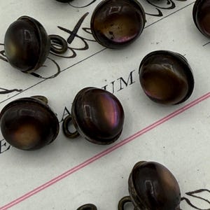 May include: A collection of antique, dark brown buttons with iridescent highlights. Each button has a rounded, domed shape with a metal loop on the back for sewing. The buttons are scattered on a vintage paper background.