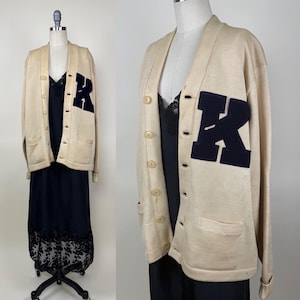 Peut inclure: Cardigan crème avec un écusson "R" en feutre bleu marine sur la poitrine gauche. Le pull a un col en V, une fermeture boutonnée et deux poches avant. Le pull est présenté sur un mannequin.