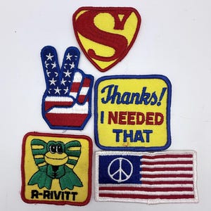 Op de afbeelding: Een verzameling van vijf geborduurde patches, waaronder een rood en geel S-schild, een handgebaar met een vredesteken in de kleuren van de Amerikaanse vlag, een geel vierkant met de tekst Thanks! I NEEDED THAT, een kikker met de tekst R-RIVITT en een Amerikaanse vlag met een vredesteken.