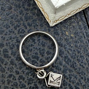 May include: A silver ring with a small square charm. The charm has a letter "K" design. The ring is on a dark textured surface, with a white and gold-trimmed frame in the background.