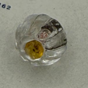 May include: A clear, round glass bead with a yellow center and a swirl design. The bead has a slightly irregular shape and appears to be handmade. The bead is a decorative item.