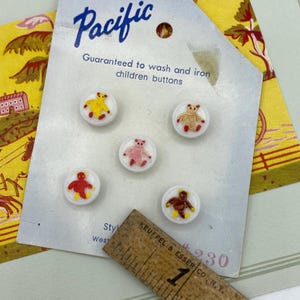 May include: A card of Pacific brand children's buttons. The card reads "Guaranteed to wash and iron children buttons." Each button is white with a colorful teddy bear design. A wooden ruler is in the foreground.