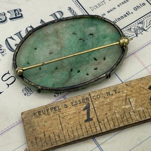 May include: A green jade oval brooch with a silver setting and a gold pin. The brooch is set against a white and blue lined paper background with a ruler that reads "KEUFFEL & ESSER CO., N.Y."