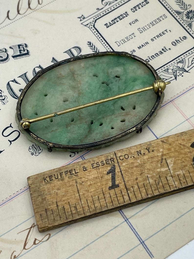 May include: A green jade oval brooch with a silver setting and a gold pin. The brooch is set against a white and blue lined paper background with a ruler that reads "KEUFFEL & ESSER CO., N.Y."