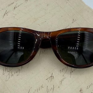 May include: Brown tortoiseshell sunglasses with dark lenses. The classic frames have a rectangular shape and are set against a vintage paper background. The sunglasses have a timeless design.
