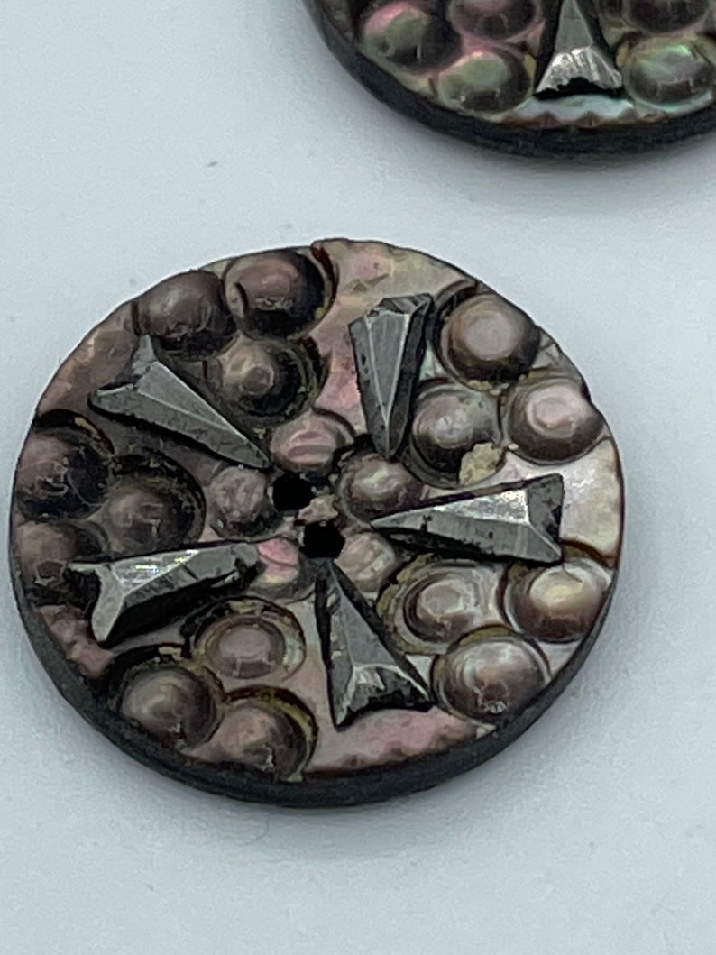 May include: A round, dark brown, iridescent button with a raised, textured design of circles and five silver-colored arrowheads.