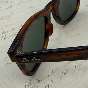 May include: Close-up of Ray-Ban sunglasses with a tortoiseshell frame. The sunglasses have dark green lenses and the Ray-Ban logo is visible on the arm. The glasses are resting on a piece of paper with handwritten script.